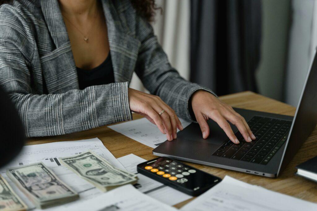 business-money-calculations A small business owner has their laptop, a calculator, and stacks of money in front of them, meant to signify doing business financial calculations.