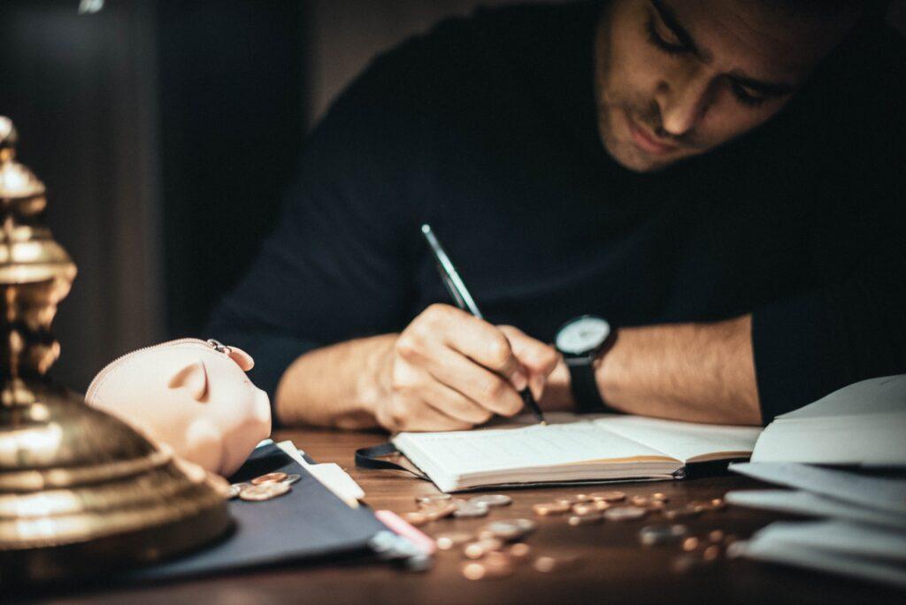 A small business owner plans their business's financial future, shown by them writing in a journal with a piggy bank and coins on the desk in front of them.