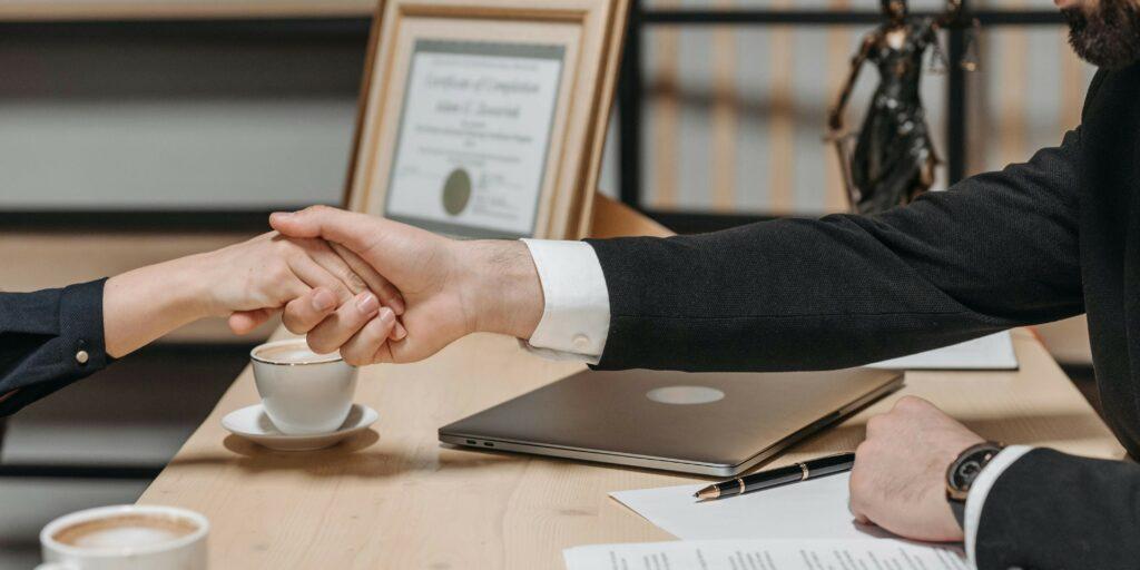 business-agreement-handshake-3 Two businesspeople shake hands at a meeting with a contract and pen on the table between them, signifying a business agreement.
